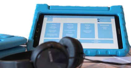 Das LSI.J Sprachtest-Tablet steht in seiner charakteristischen blauen Hülle auf einem Tisch. Daneben liegen Kopfhörer.